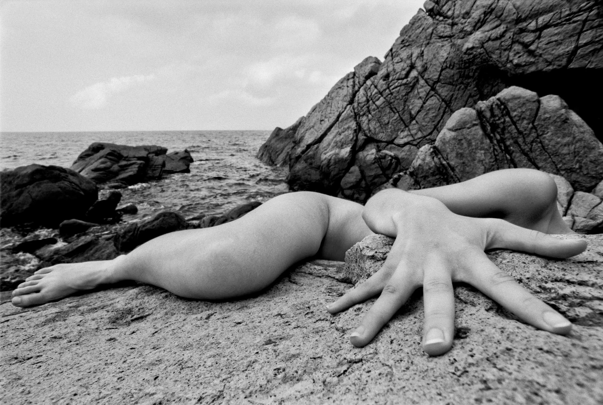 Black and white fine art nude study of a reclining female figure on coastal rocks, with an outstretched hand reaching into the foreground.