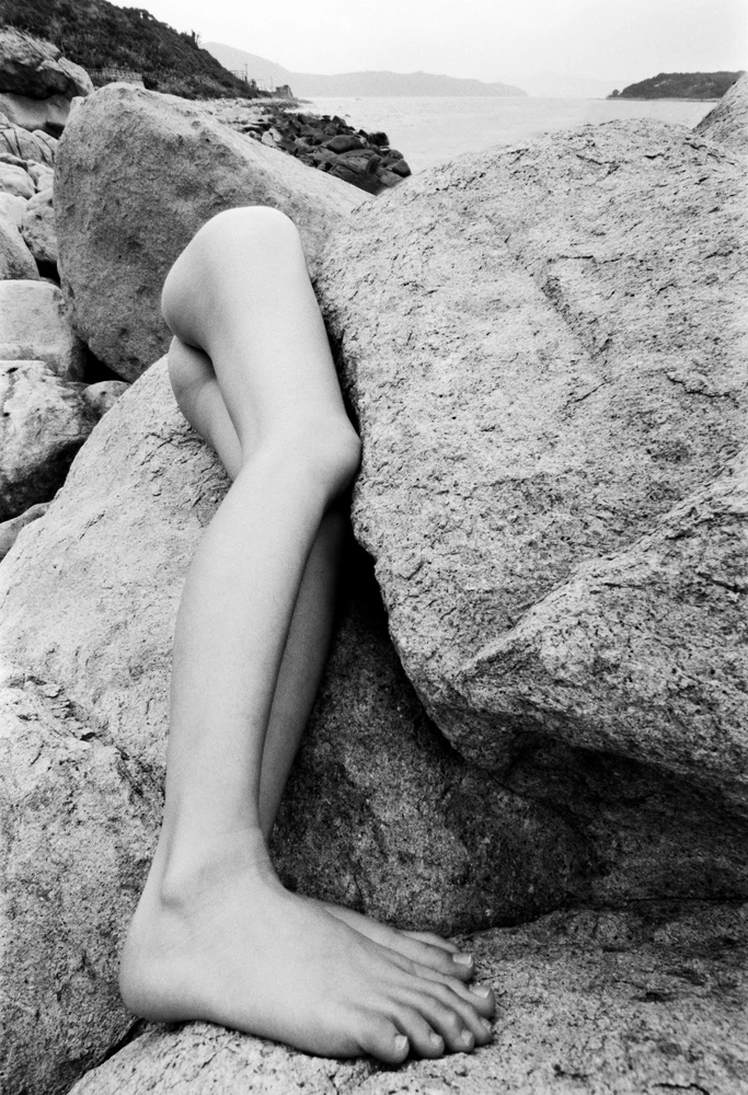 Black and white fine art nude photograph of a reclining female figure on coastal rocks, with elongated, distorted legs dominating the foreground against the sea and distant mountains.