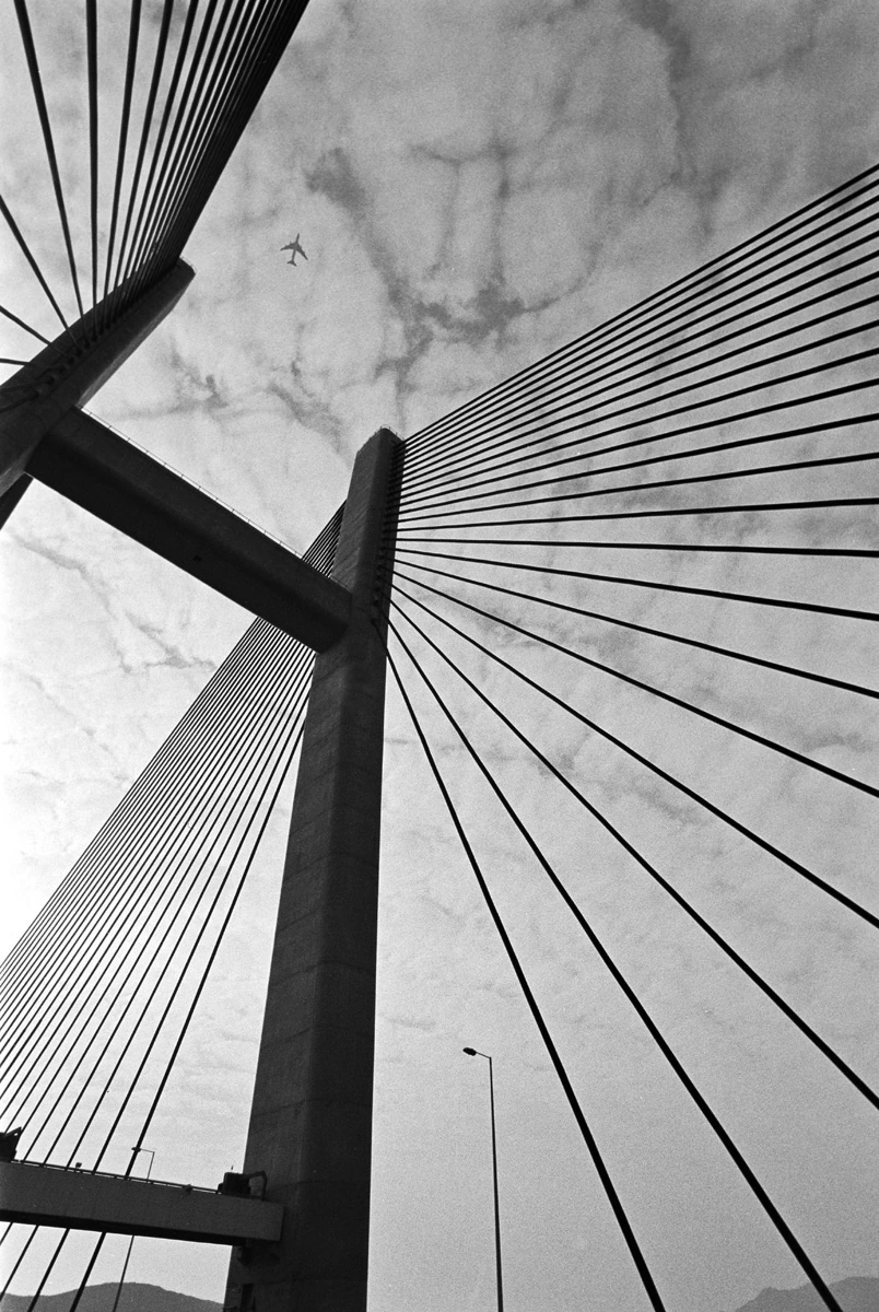 Black and white fine art photograph of a cable bridge in Hong Kong featuring strong architectural geometry, with an aircraft flying overhead.